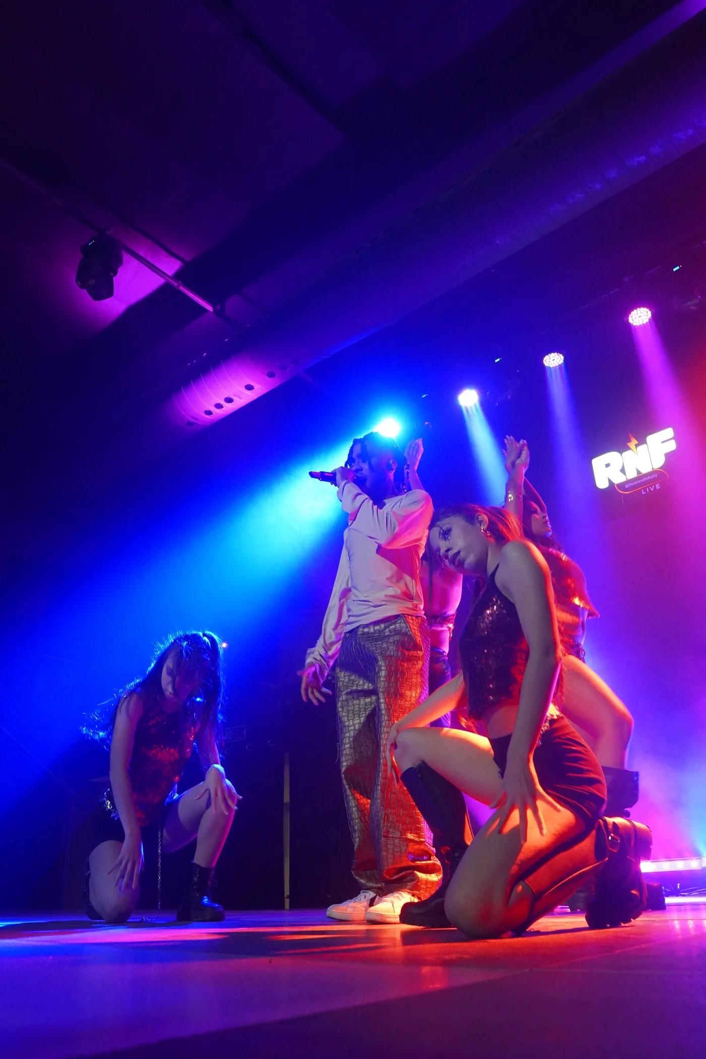 Reny cantando acompañado de bailarinas bajo luces azules y violetas